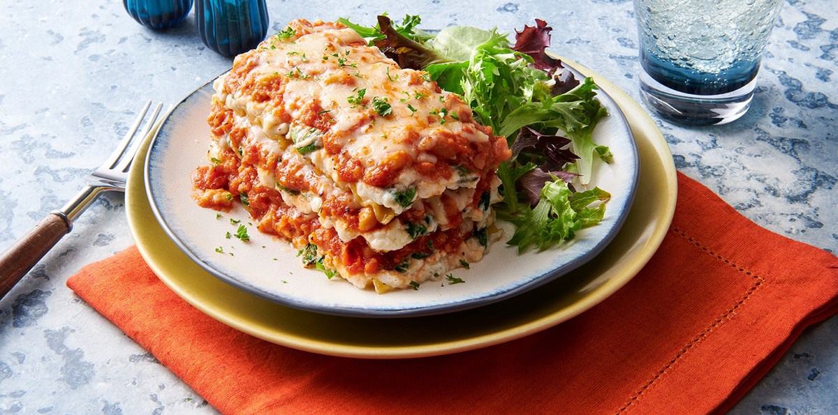 Garbanzo-Spin Ricotta Lasagna is plated next to a side of greens. Next to the plate is salt and pepper, a beverage, and an orange napkin. 